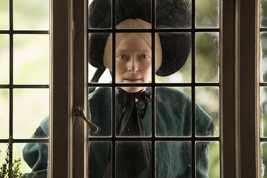 Tilda Swinton stands with serious expression looking into house windows and wears Victoria era black bonnet and dark green dress