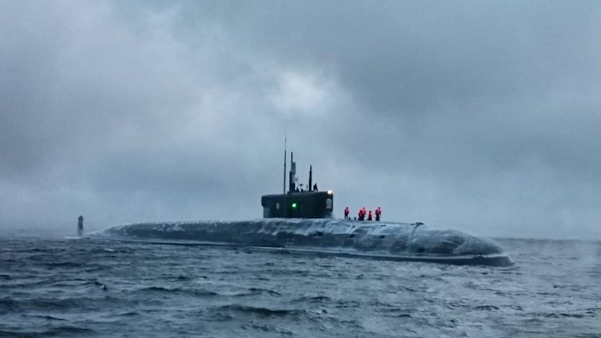 In cold overcast weather, you view a submarine in icy waters with personnel in red hi-vis on top of it.