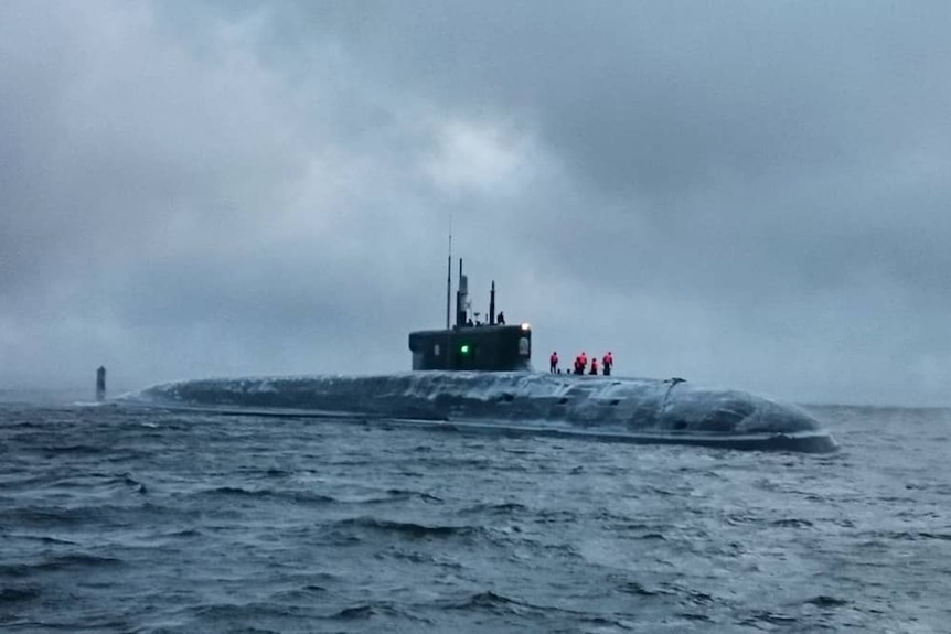 In cold overcast weather, you view a submarine in icy waters with personnel in red hi-vis on top of it.