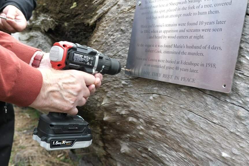 A man wearing an orange jumper holding a power drill drills a hole through a plaque that says rest in peace on a log.