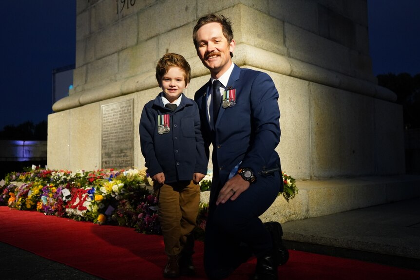Father and son in uniform stand in front of a war memorial 