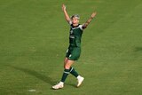 A soccer player wearing green and white holds her arms in the air with a surprised look on her face