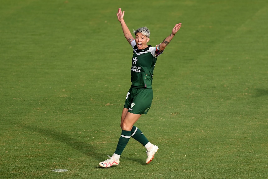 A soccer player wearing green and white holds her arms in the air with a surprised look on her face