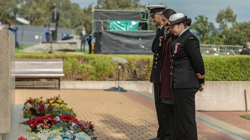 Three people looking reflectively at wreaths laid on monument, two are in uniform