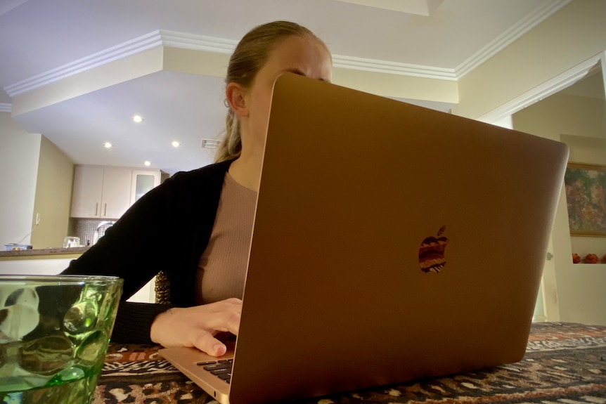 A young woman is using a laptop at the kitchen table, her face is hidden behind the screen.