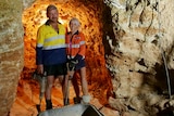 Gregg and Lynette in high visibility shirts holding tools, underground, light behind.