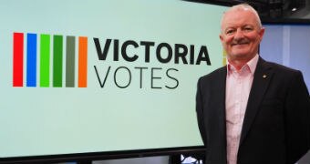 Antony Green stands in front of a screen which displays a Victoria Votes logo.