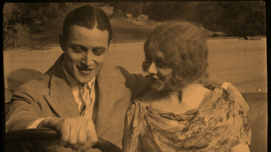 A grainy image from a 1929 film showing a man in a suit driving a car with his arm around a woman in a beautiful dress.
