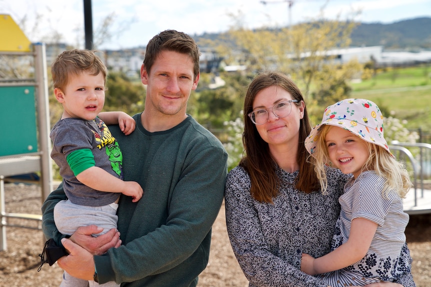 Married couple Courtney and Jason smile at the camera, holding one young child each, at a playground.