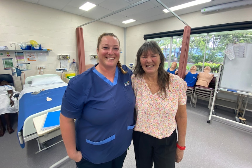 A woman in blue nursing scrubs next to an older woman in a university training room set up to look like a hospital ward.