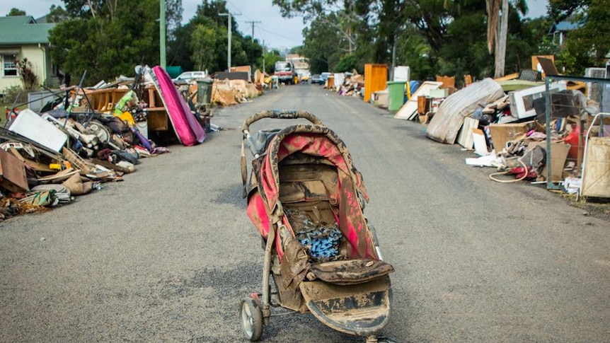 Piles of household items line a suburban road, a child's pram covered in mud is centre frame.
