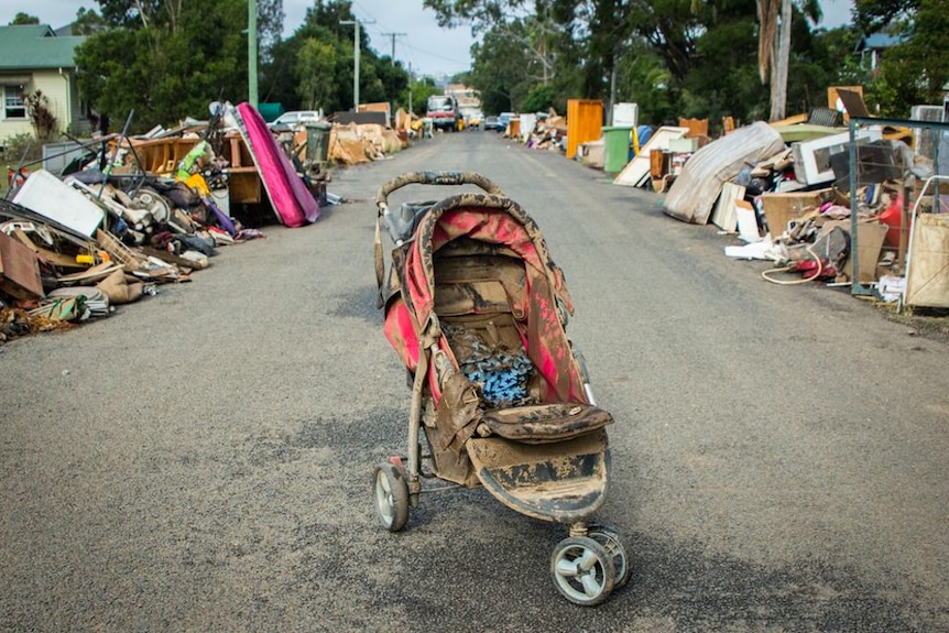 Piles of household items line a suburban road, a child's pram covered in mud is centre frame.