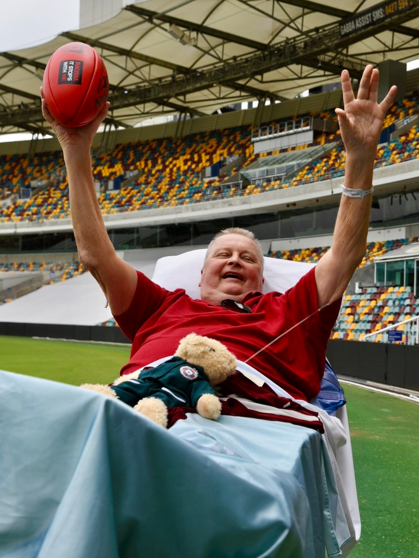 A man in a hospital bed holds up a football at a stadium.