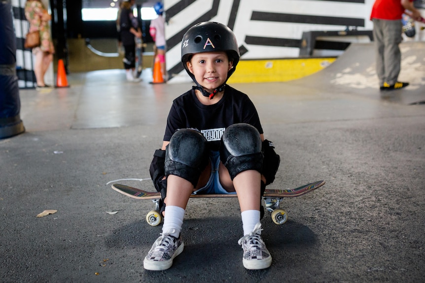 A young girl in all black sits atop her skateboard. She is smiling. Her helmet as an A on it.