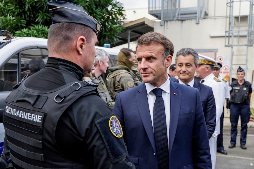 A police officer in uniform shakes the hand of Emmanuel Macron, who is in a blue suit and tie looking serious