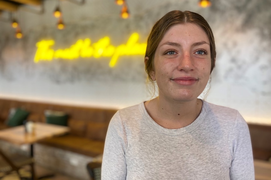 Femme avec une chemise blanche souriante debout devant une enseigne au néon jaune