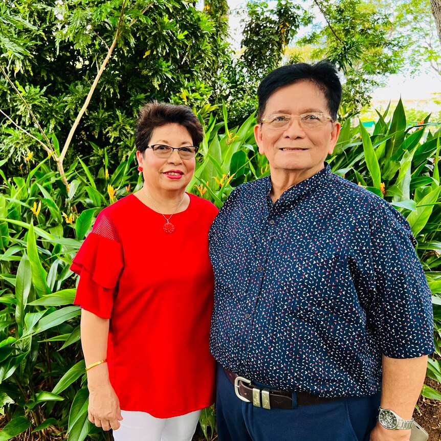 man in blue shirt and woman in red blouse stand next to each other in front of a green garden