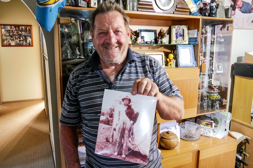 A man wearing a striped blue shirt holds up an old sepia photo of a beekeeper.