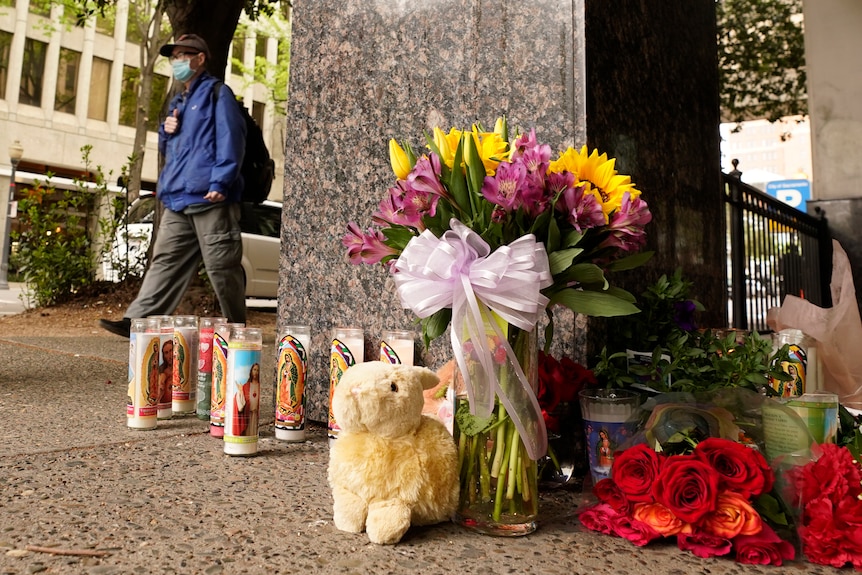 A small memorial of candles, flowers and a plush rabbit toy next to a concrete pillar, as a person walks past.