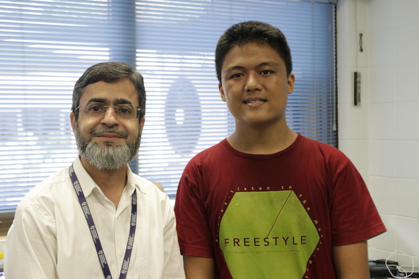 A male university lecturer and a teenage boy, standing next to each other and smiling at the camera, inside a classroom.