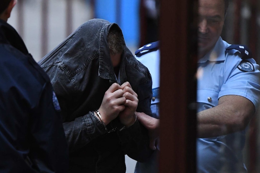 A man in handcuffs with a shirt over his head is flanked by two male police officers, one of whom has a hold of his arm.