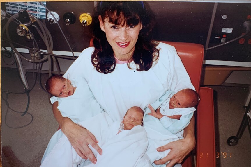 A mum with brown hair sitting in a chair holding three babies. She has a white hospital gown on. 