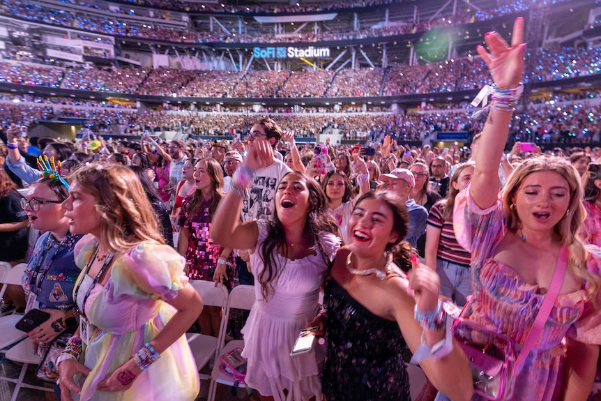 A crowd of screaming young women dance with their hands up as they're bathed in pink light.