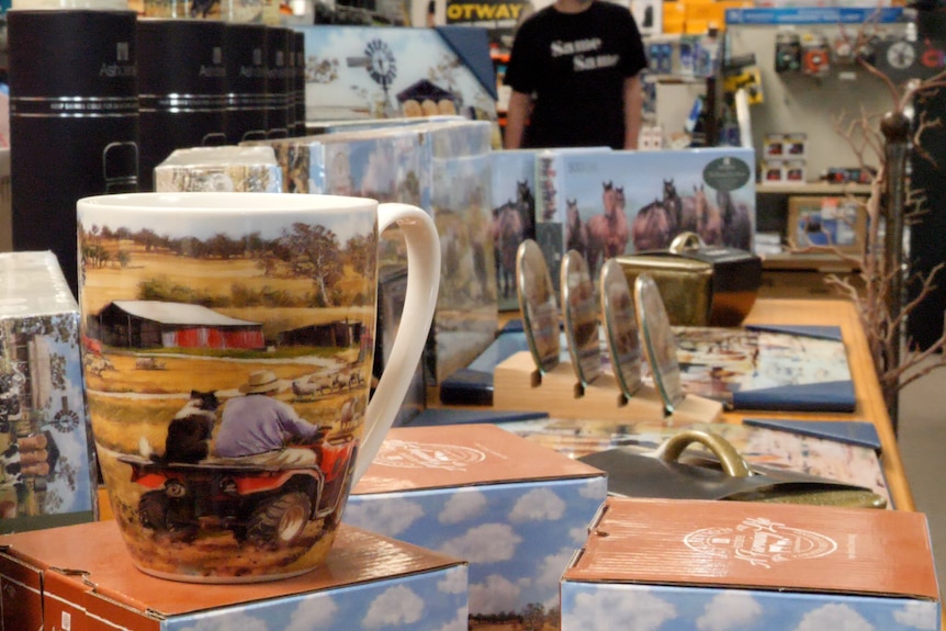 Mugs printed with a painting of a man on a tractor with a border collie dog in a hardware store.