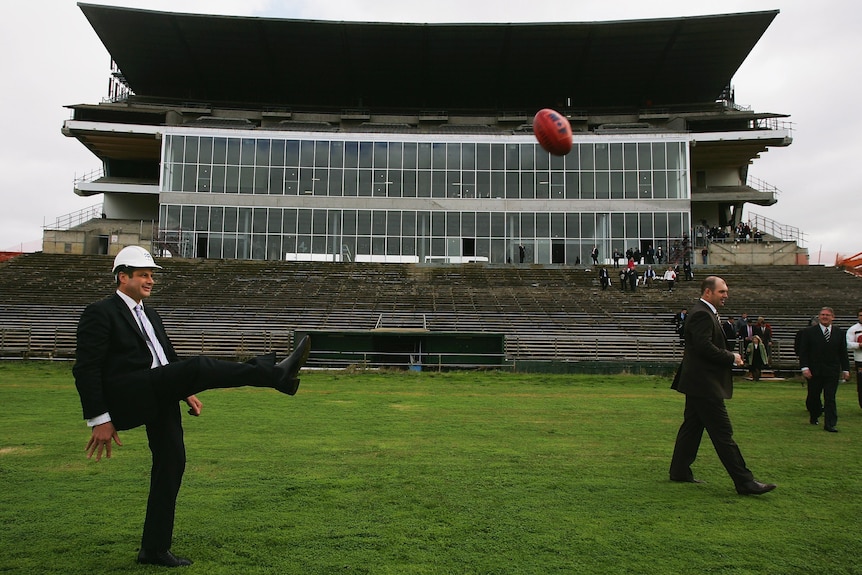 Steve Bracks wearing a hard hat kicks a football on Waverley Park.