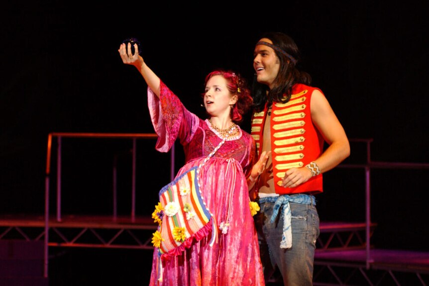 A white woman with red hair wearing a medieval-styled pink gown stands with a white man with long dark hair and red and vest.