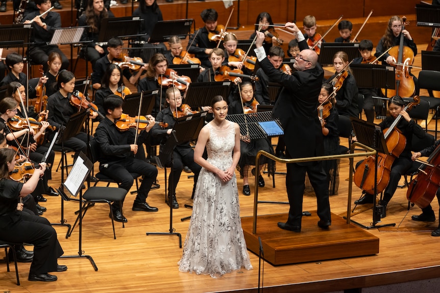 A woman stands in a long dress in the middle of an orchestra and next to the conductor.