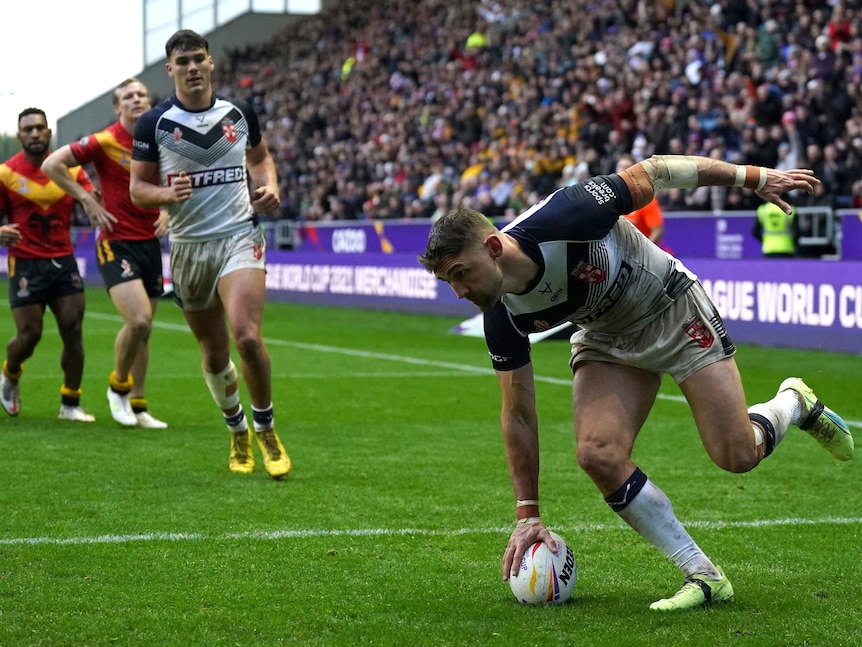 An England rugby league player leans in to touch the ball down for a try as England and PNG players watch.