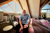Bald man sitting on bed, in front of two windows in a teenage girl's bedroom
