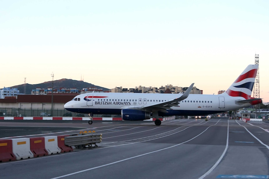 A large British Airways plane sits on an airport runway. A mountain can be seen in the distant background.