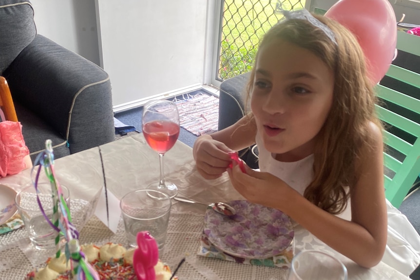 a girl sits at a table with a cake in front of her. it has a candle in the shape of an eight