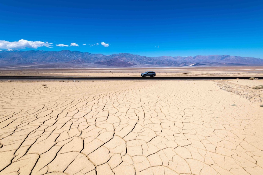 Crack dry ground in the foreground and a blue car drives on a road.