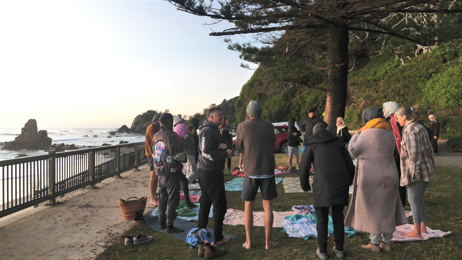 Group of 20 people stand in a circle on grass with the beach and rocks in the background 