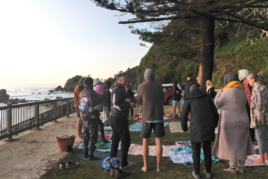 Un groupe de 20 personnes se tient en cercle sur l'herbe avec la plage et les rochers en arrière-plan 