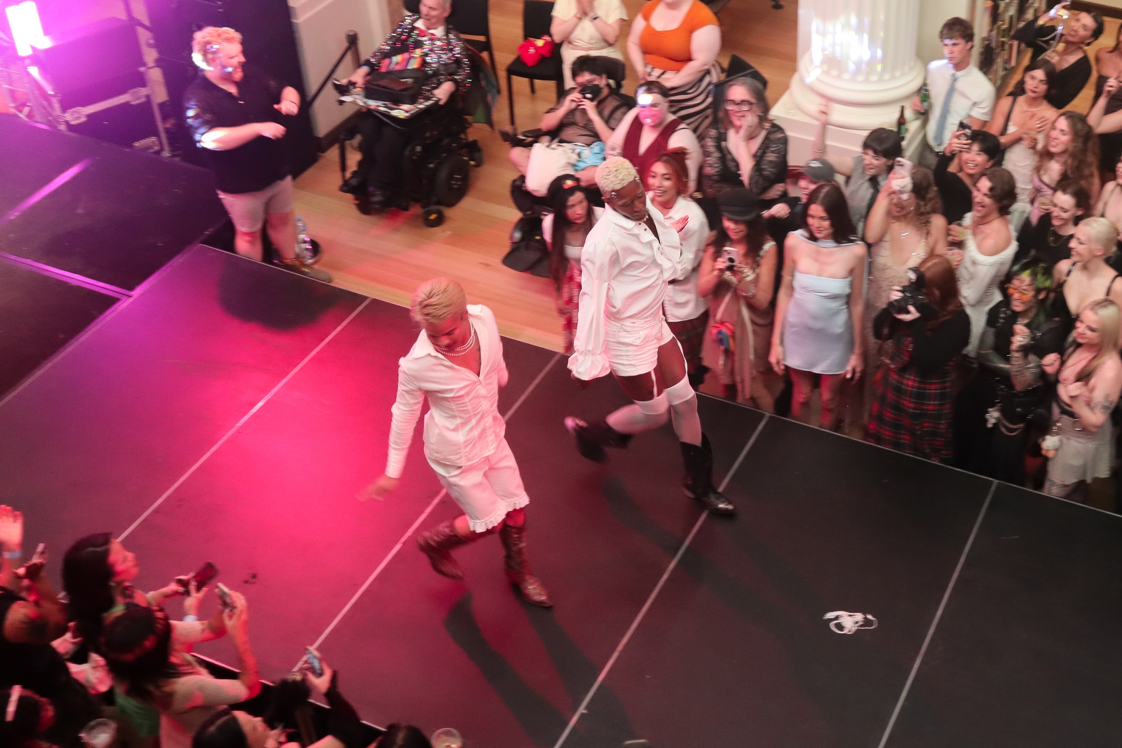 Dos personas vestidas de blanco se pavonean en el escenario frente a los demás.