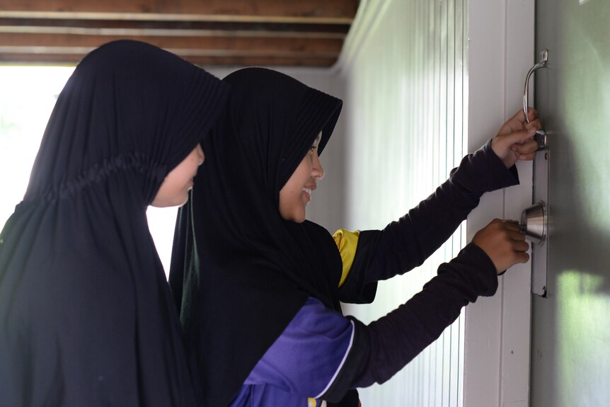Two primary school students wearing hijabs unlock a green door.