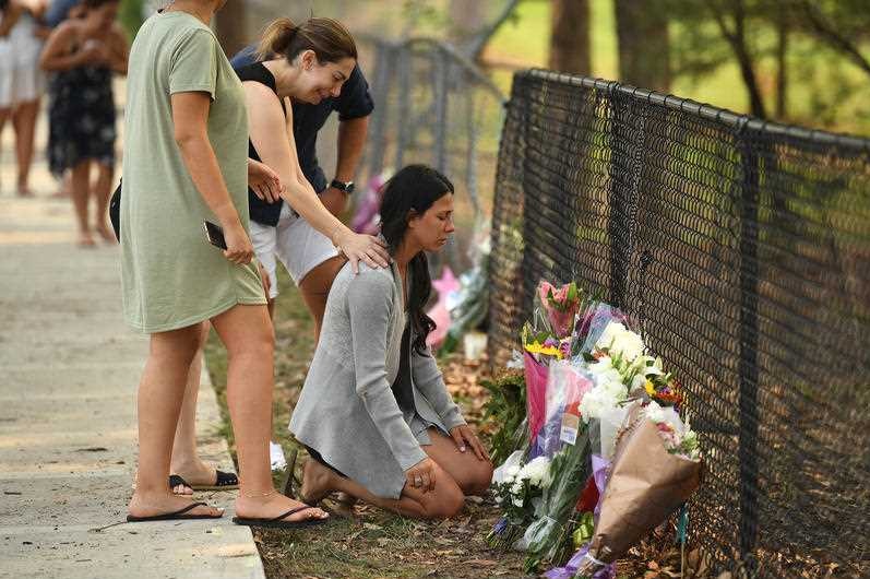 A woman in a grey cardigan kneels in front of memorial flowers.