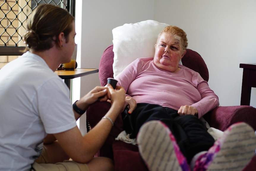 A white woman with short red hair and a bandage on her head. She's talking to her son while sitting in an arm chair