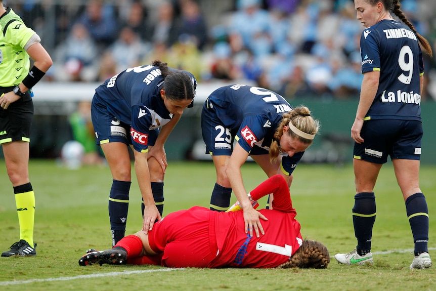 A soccer goalkeeper wearing red lays on her side as her team-mates in dark blue check on her welfare