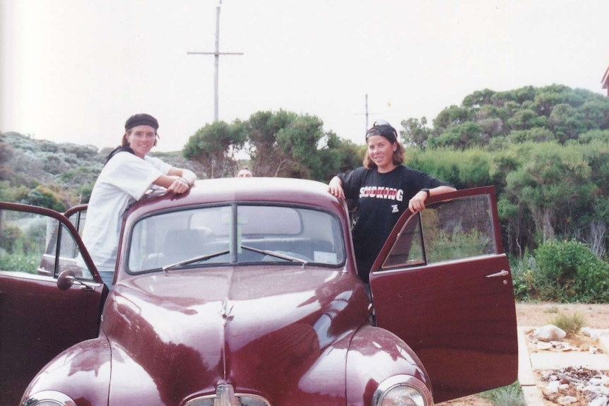 Two young women standing in the front doors of an old brown Beetle car.
