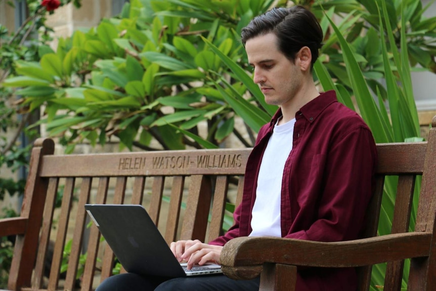 A university student sitting on a bench and working on their laptop.