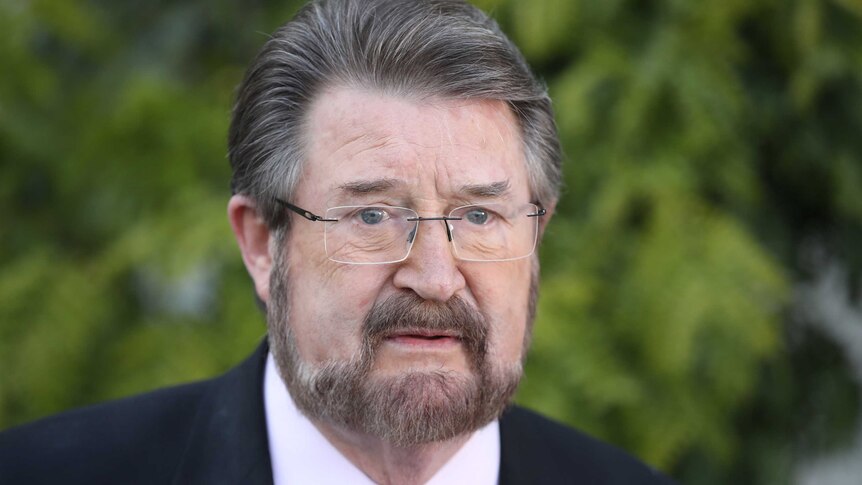 Derryn Hinch frowns while looking just past the camera. He is wearing a dark suit with a pink, red and grey patterned tie.