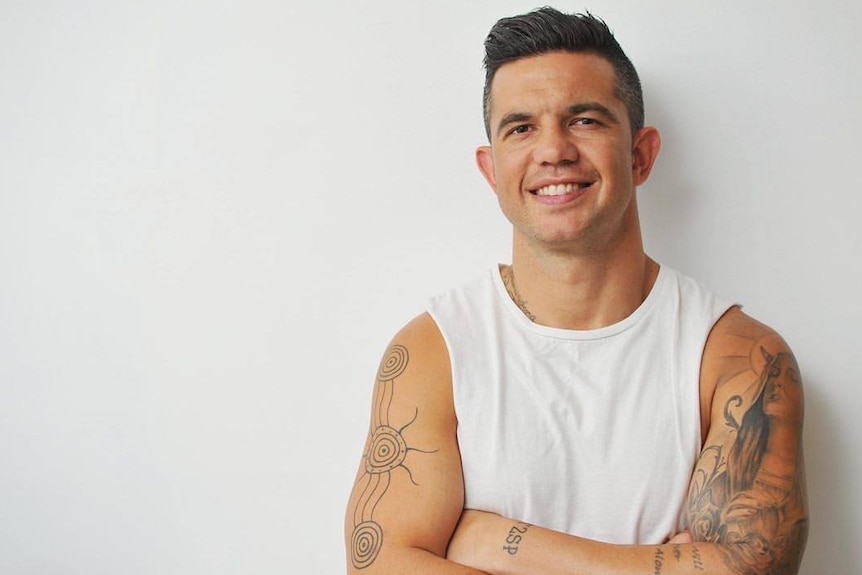 Joe Williams stands in a white singlet, smiling with his arms folded.