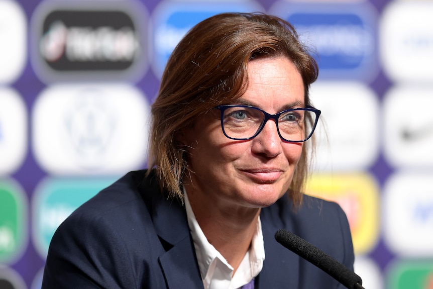 A woman wearing glasses and a jacket speaks into a microphone with a wall of sponsors behind her