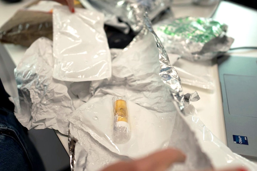 A vial in a small yellow container sits on a pile of foil, melted ice packs, and other packaging on a desk.