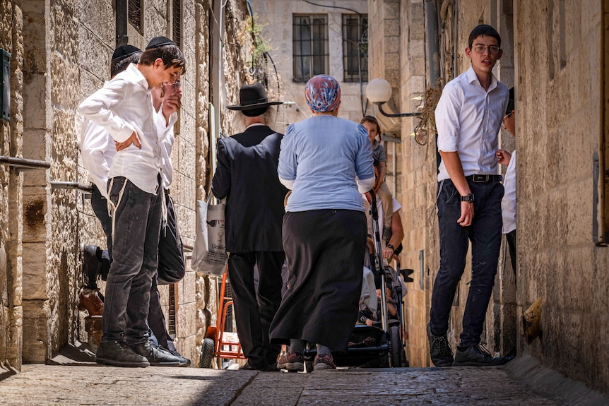 A man in traditional Orthodox garb and a woman in a long black skirt walk down a narrow Jerusalem street, past a group of boys